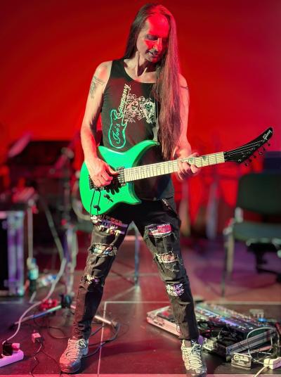 Gitarrist Joe steht auf einer Bühne in rotem Licht, er spielt seine leuchtend grüne Gitarre, er hat langes, dunkles Haar und ein Shirt seiner Band an