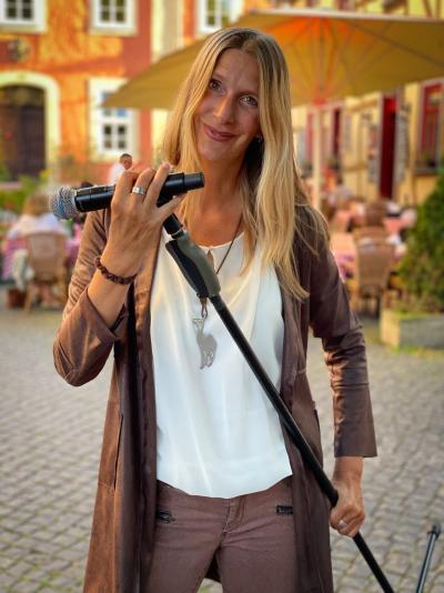 Sängerin Stephanie steht mit ihrem Mikrofon im Mikrofonständer in der Hand auf einem Platz, sie hat lange, blonde Haare, trägt ein weißes Shirt und darüber eine braune, längere Jacke